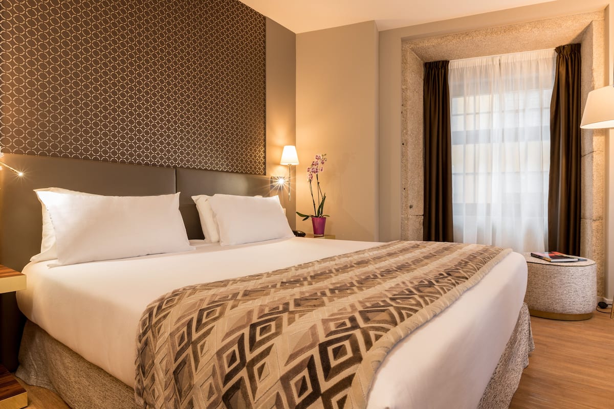 Un lit bien fait, recouvert d’une couvre-lit à motifs, avec une tête de lit au design géométrique, une lampe de chevet et une fenêtre avec des rideaux.