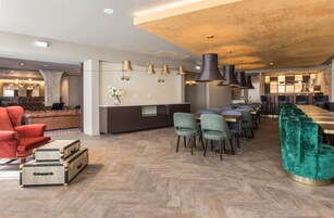 Spaziosa hall d'albergo con pavimento in parquet a spina di pesce, un'area bar centrale e un salotto con poltrone confortevoli e un divano rosso.