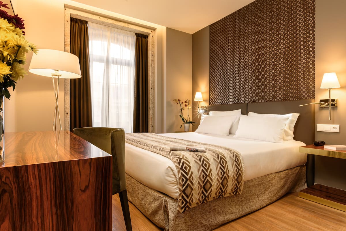 Une chambre d’hôtel avec un grand lit, un bureau avec une lampe, une chaise et une fenêtre avec des rideaux.