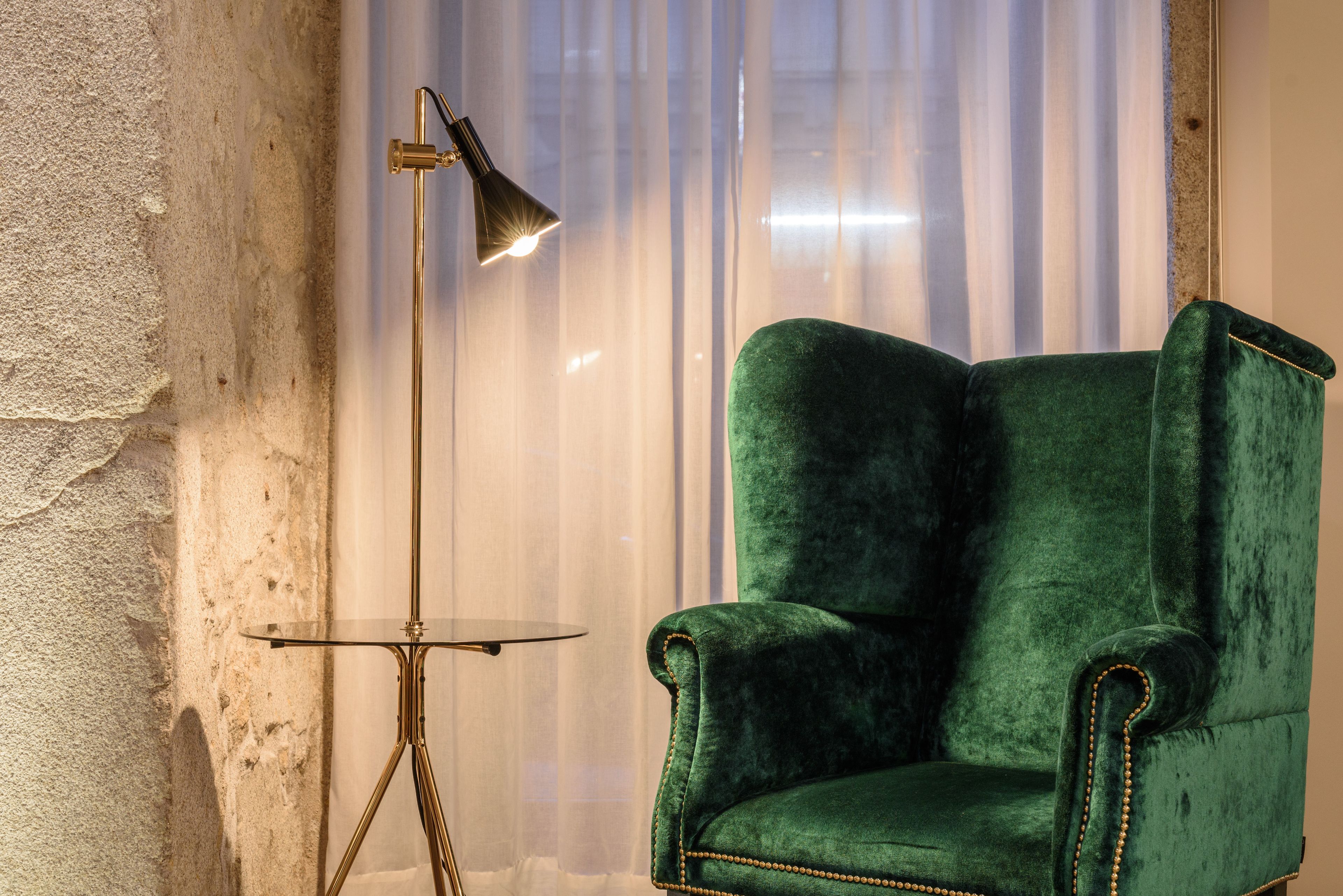 A green velvet armchair with a gold-trimmed armrest, positioned next to a floor lamp with a gold base and a black shade.