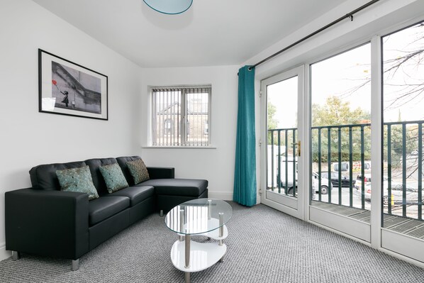 A modern living room with a black sectional sofa, a glass coffee table, and a large sliding glass door leading to an outdoor balcony.