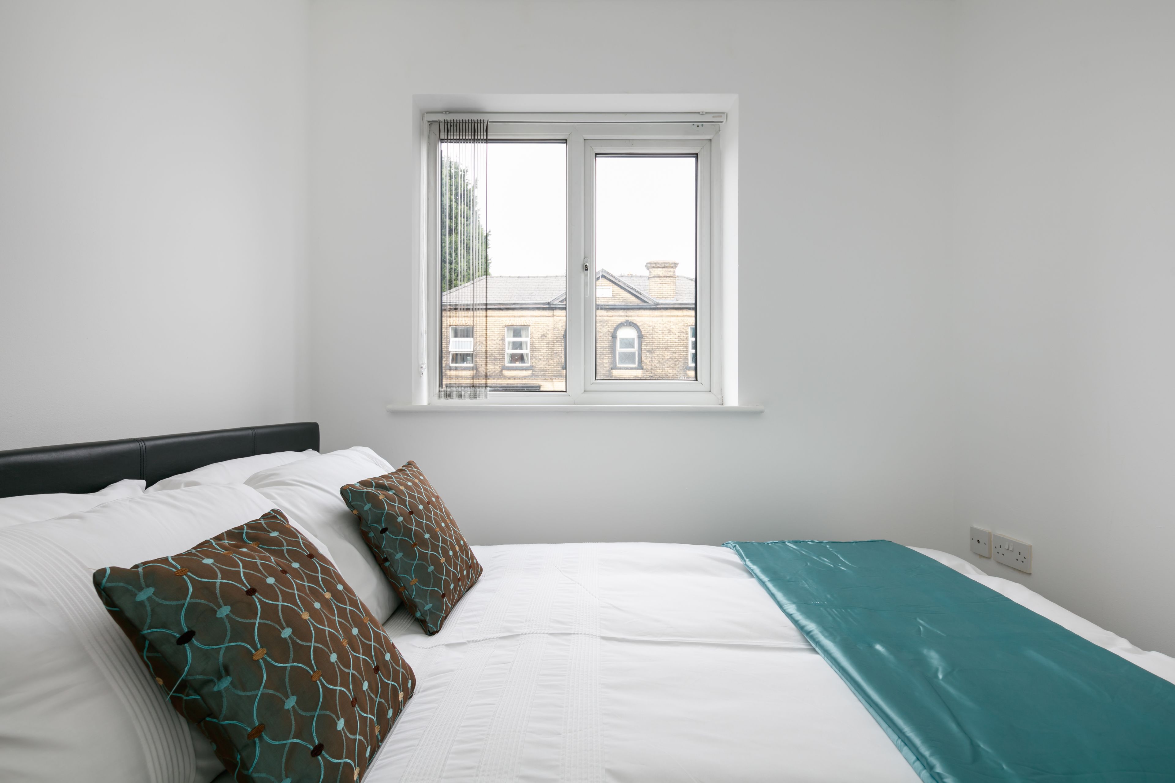 A bedroom with a bed, two patterned pillows, a teal bedspread, and a window with a view of buildings.