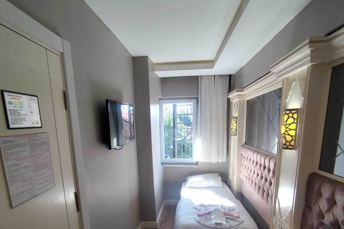 Ein kleines Hotelzimmer mit einem Bett, einem Schreibtisch, einem Sessel, einem Fernseher und einem fenster mit Vorhängen.