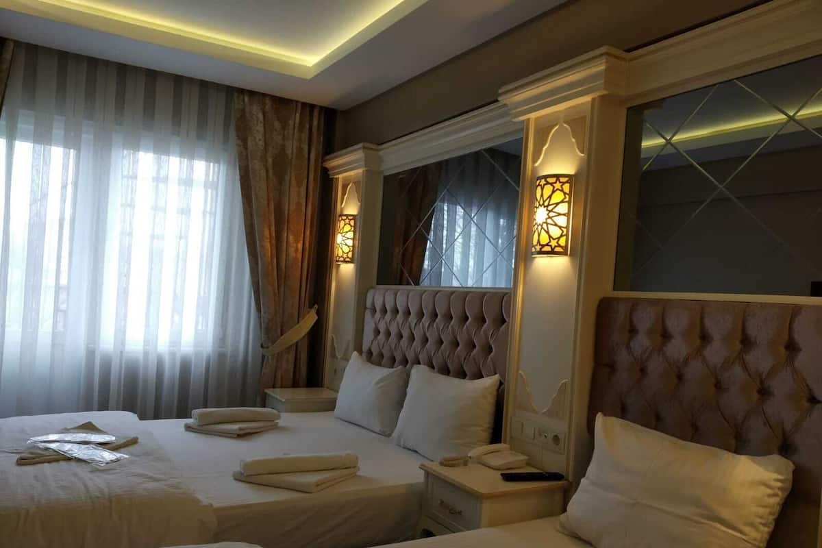 Ein Hotelzimmer mit zwei Betten, einem großen Fenster mit durchsichtigen Vorhängen, einem gepolsterten Kopfteil und zwei Wandleuchten.
