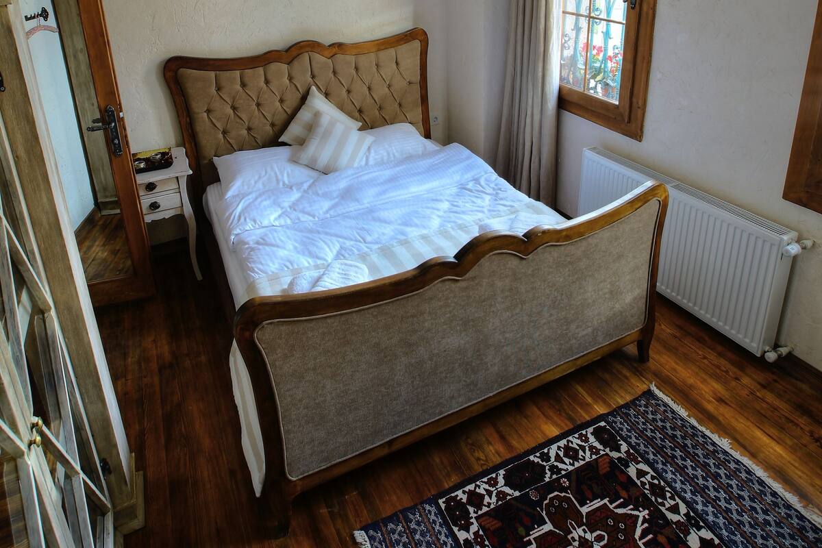 A bedroom with a bed, wooden floor, a ceiling fan, a window with curtains, and a radiator.