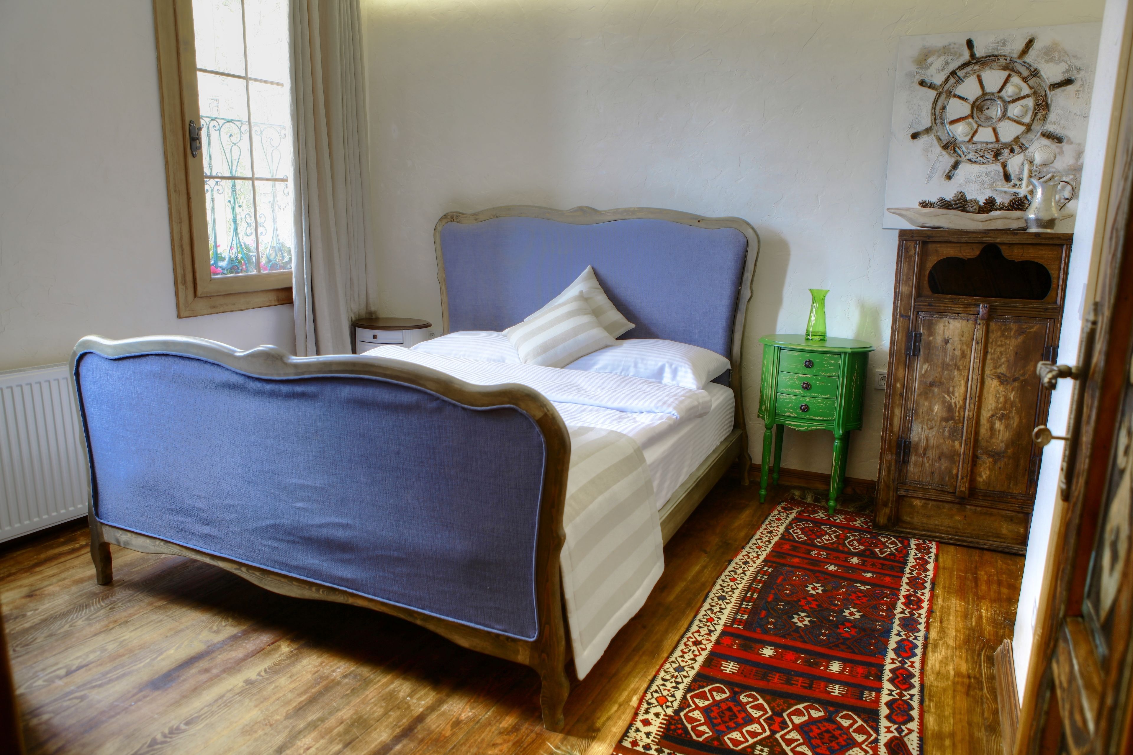 A bedroom with a bed, a wooden cabinet, a green nightstand, a ship's wheel decoration, and a patterned rug.