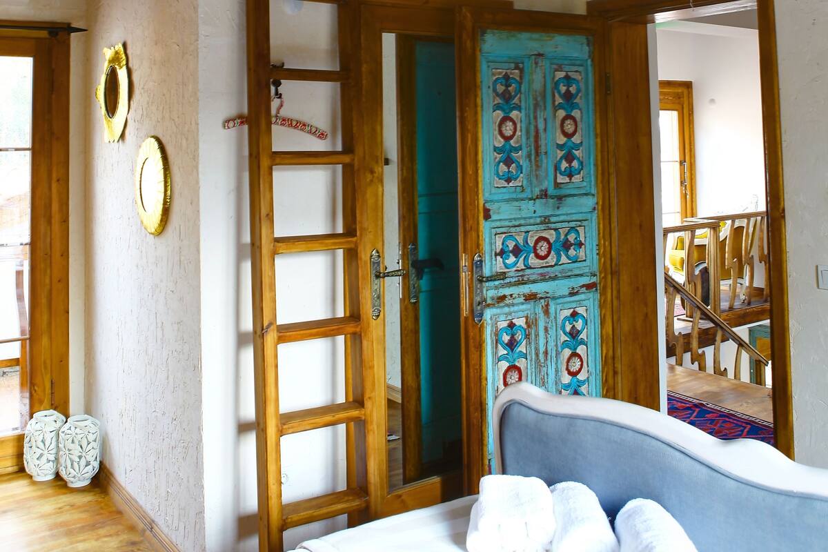 A bedroom with a wooden ceiling, a bed with white bedding, a blue headboard, and a ladder leading to a door with a blue and red pattern.