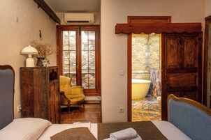Una habitación con cama, una silla, un armario de madera y un baño visible a través de una puerta de cristal.