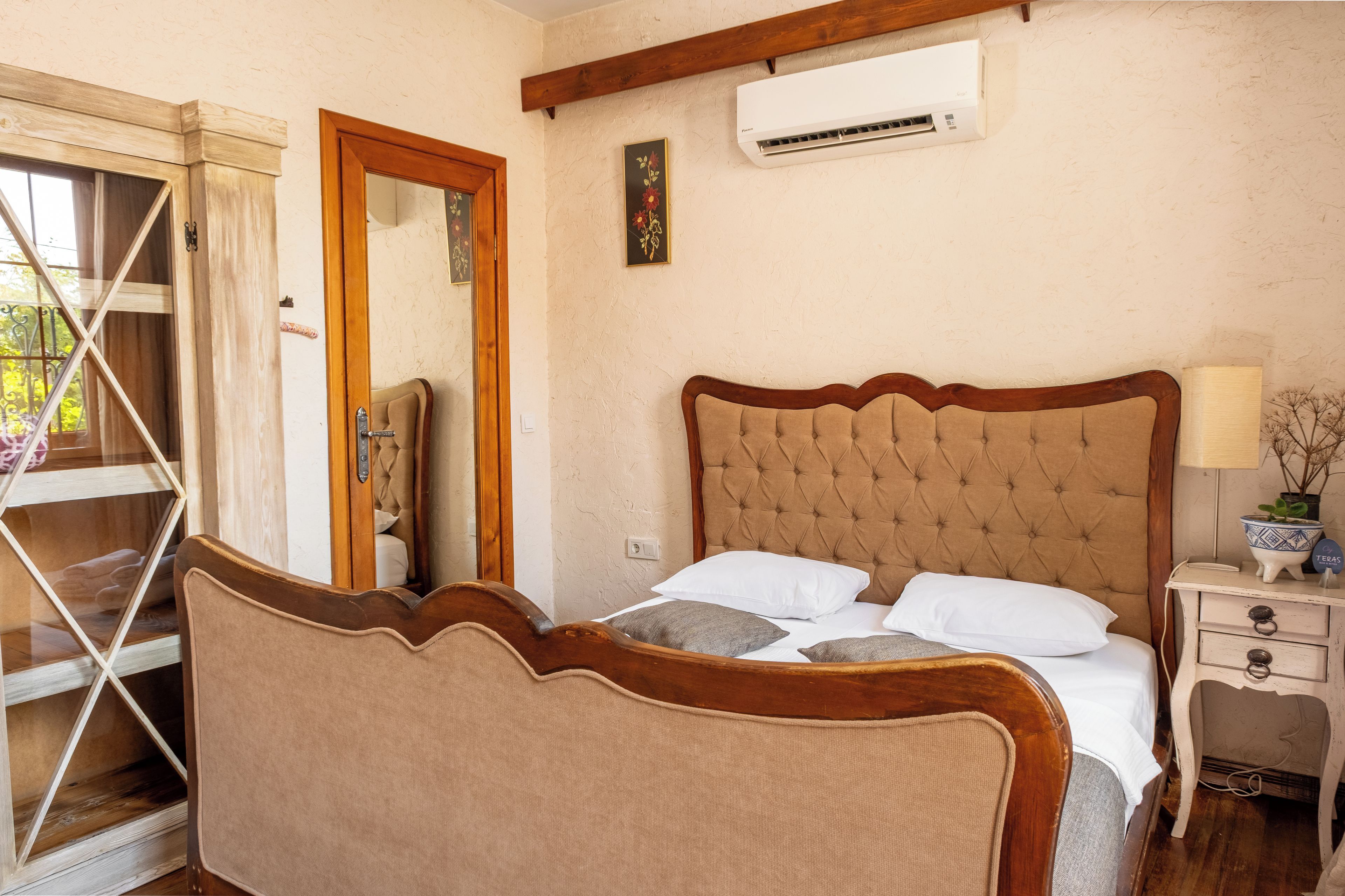A bedroom with a wooden headboard, a bed with white bedding, a nightstand with a lamp, and a window with a view of greenery.