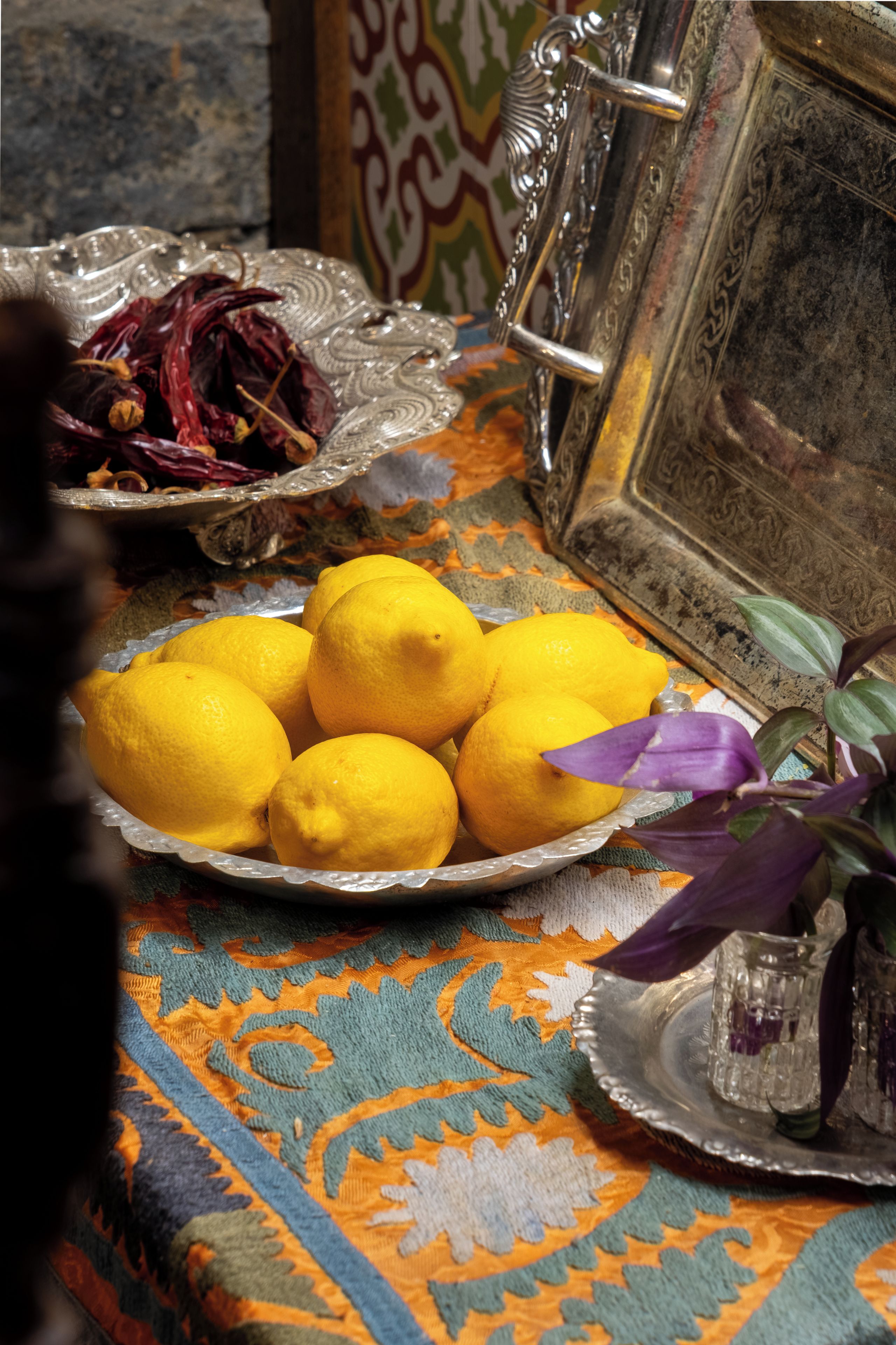 A table with a decorative plate of lemons, a glass bowl with dried flowers, and a silver tray with a glass container.