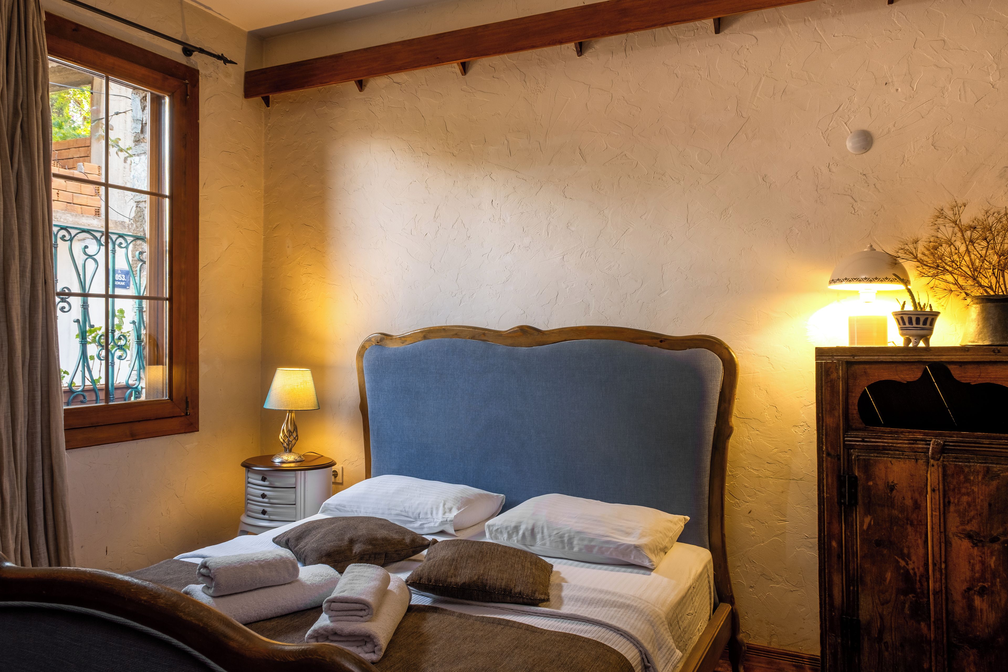 A bedroom with a bed, pillows, a bedside lamp, a wooden headboard, a nightstand, and a window with curtains.