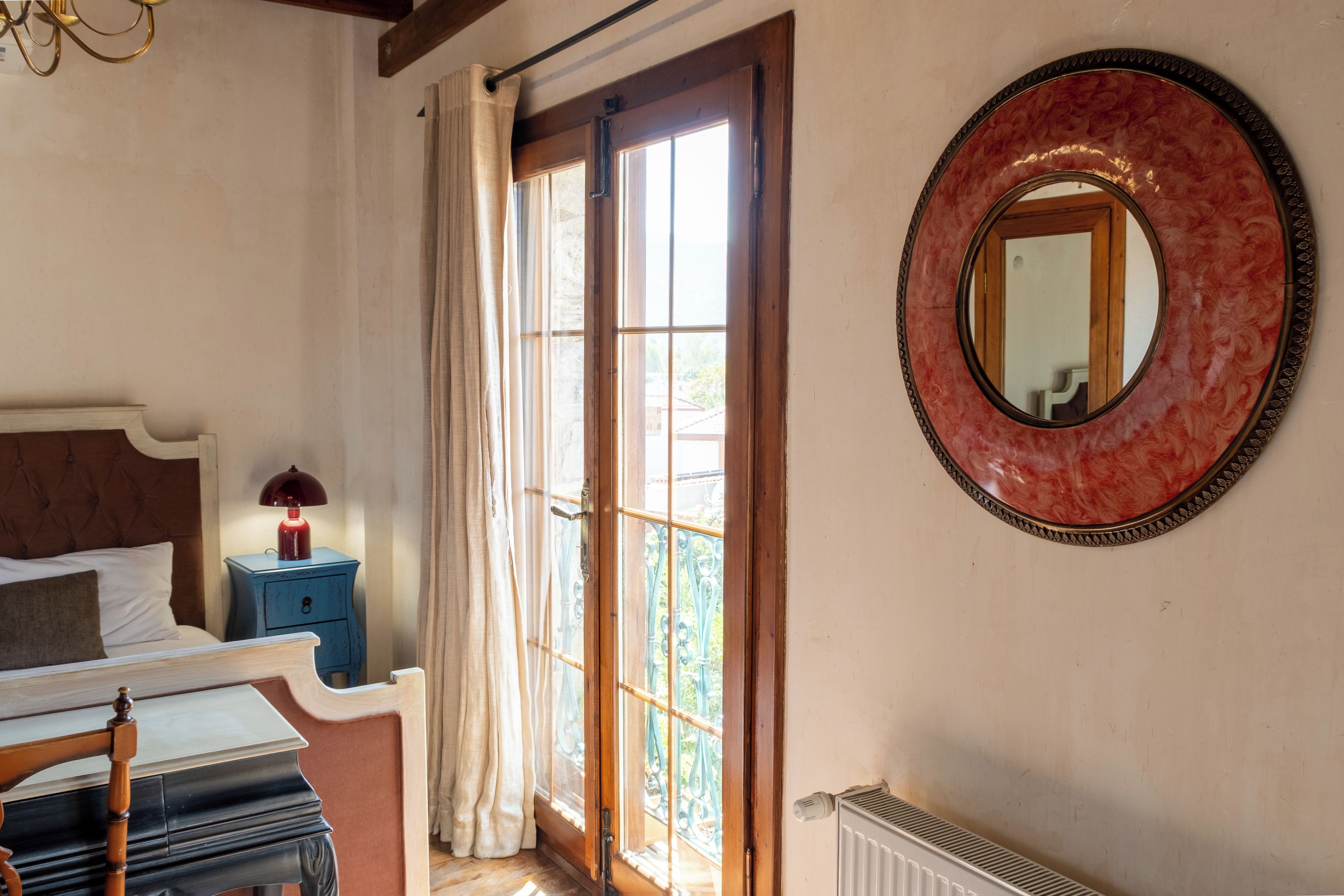 A bedroom with a bed, a nightstand, a lamp, a window with curtains, and a wall-mounted mirror.