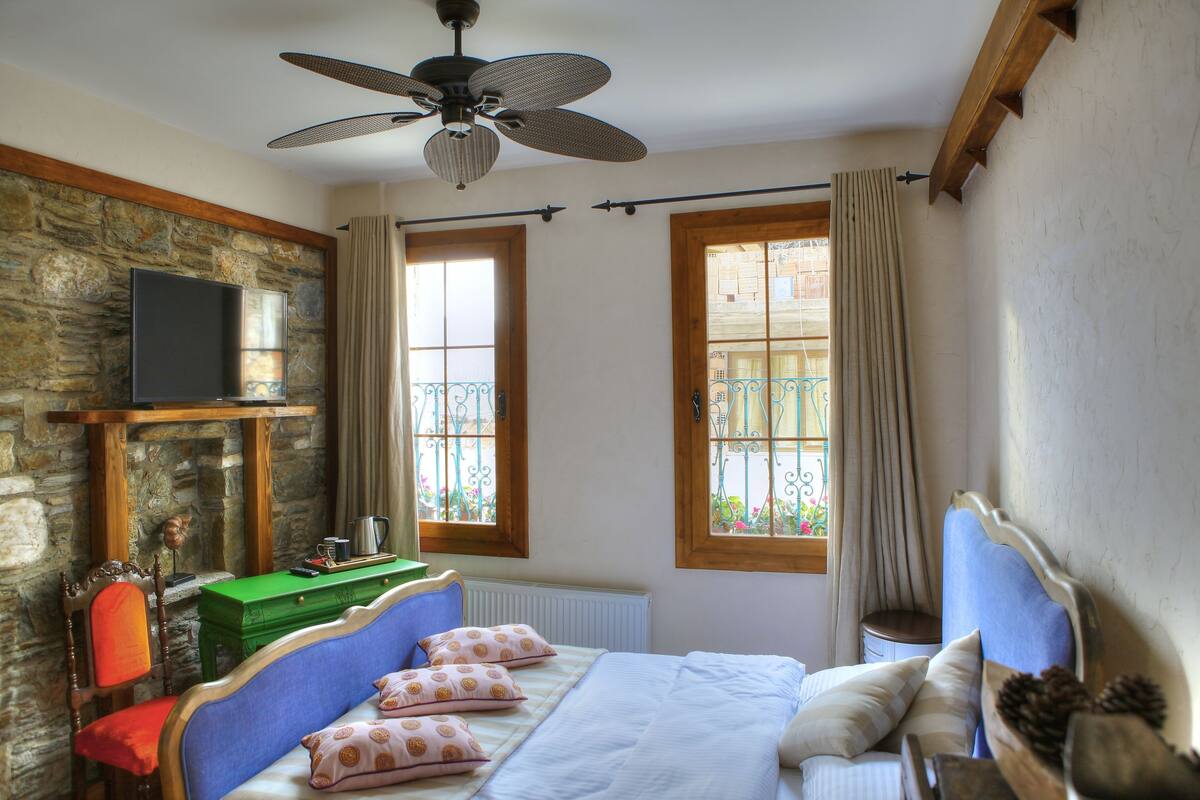 A bedroom with a stone wall, a bed with patterned pillows, a green side table, a television, and a ceiling fan.