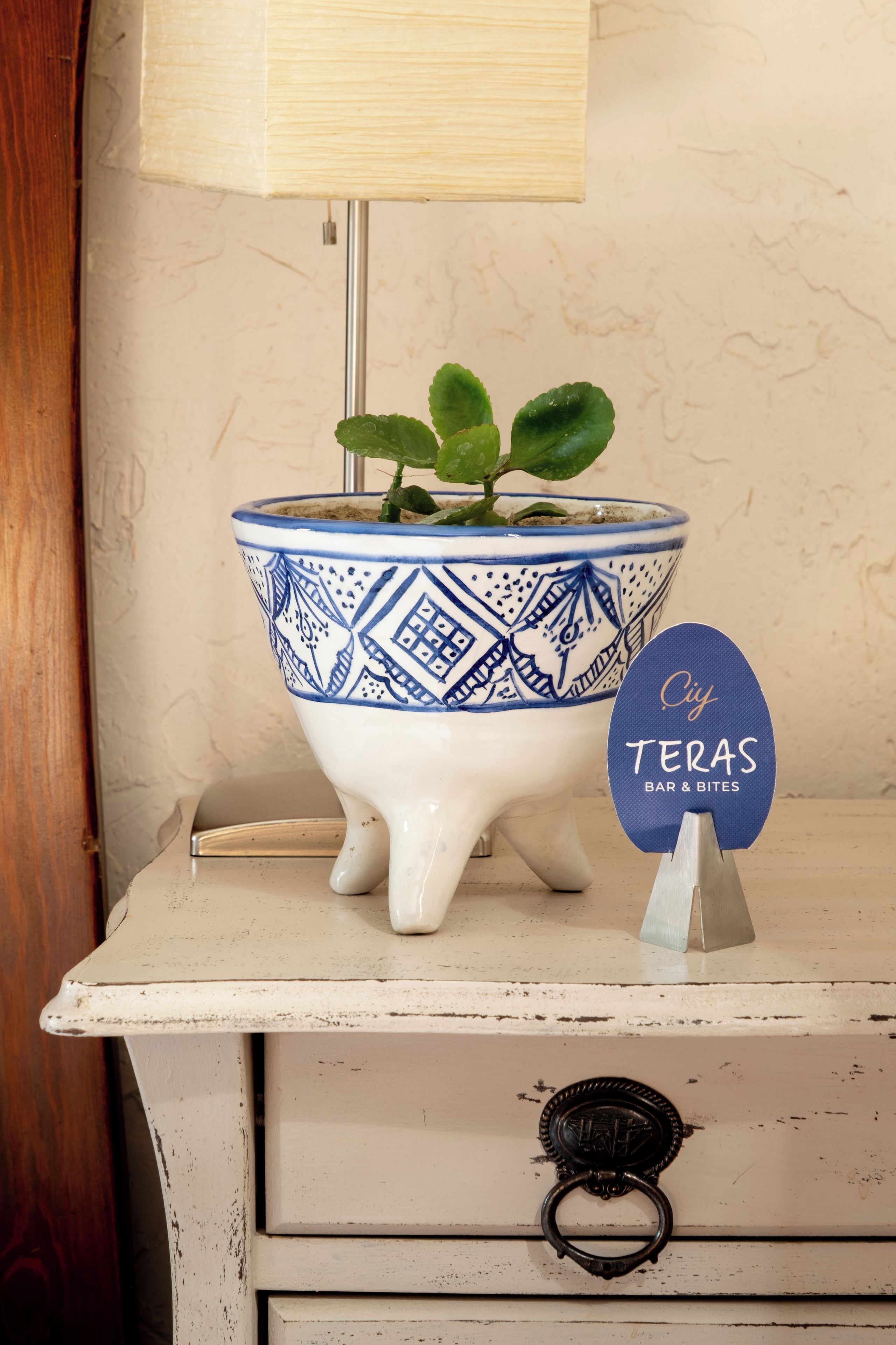 A white bedside table with a blue and white patterned ceramic pot containing a plant, a small blue sign with 'TERAS BAR & BITES' text, and a metal lamp base.