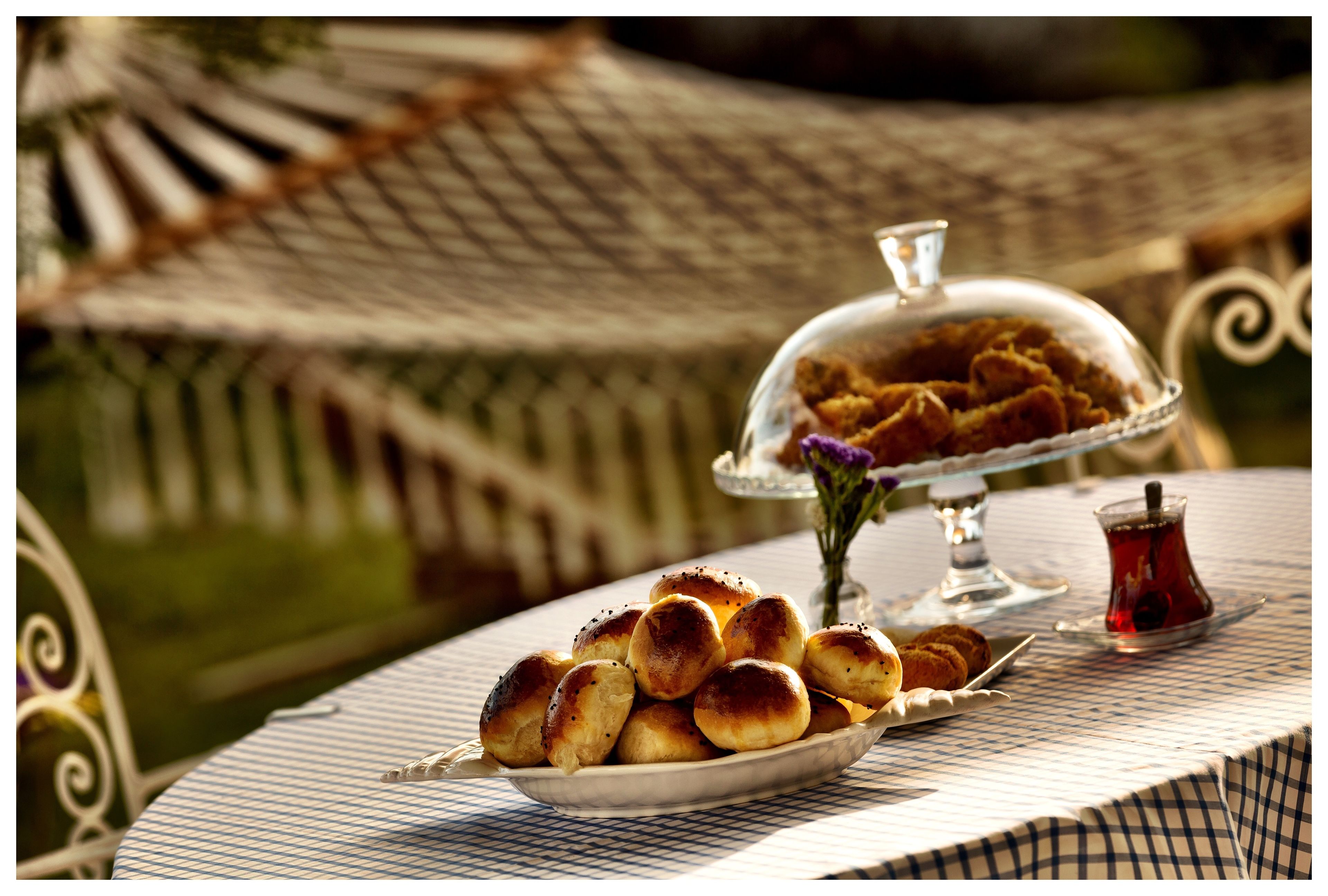 A table with a checkered tablecloth, a plate of bread rolls, a glass container with a lid, and a small glass with a red substance.