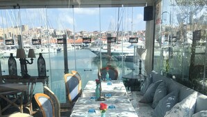 A dining area with a view of the marina, featuring tables set with blue glassware and a vase with a red flower. - Deniz Kabugu Hotel (Cesme)