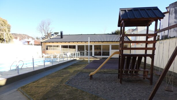 Un patio trasero con piscina, un parque infantil de madera y una zona de estar cubierta.