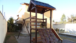 Estructura de juego de madera con un tobogán, ubicada sobre una superficie de grava en una zona residencial.