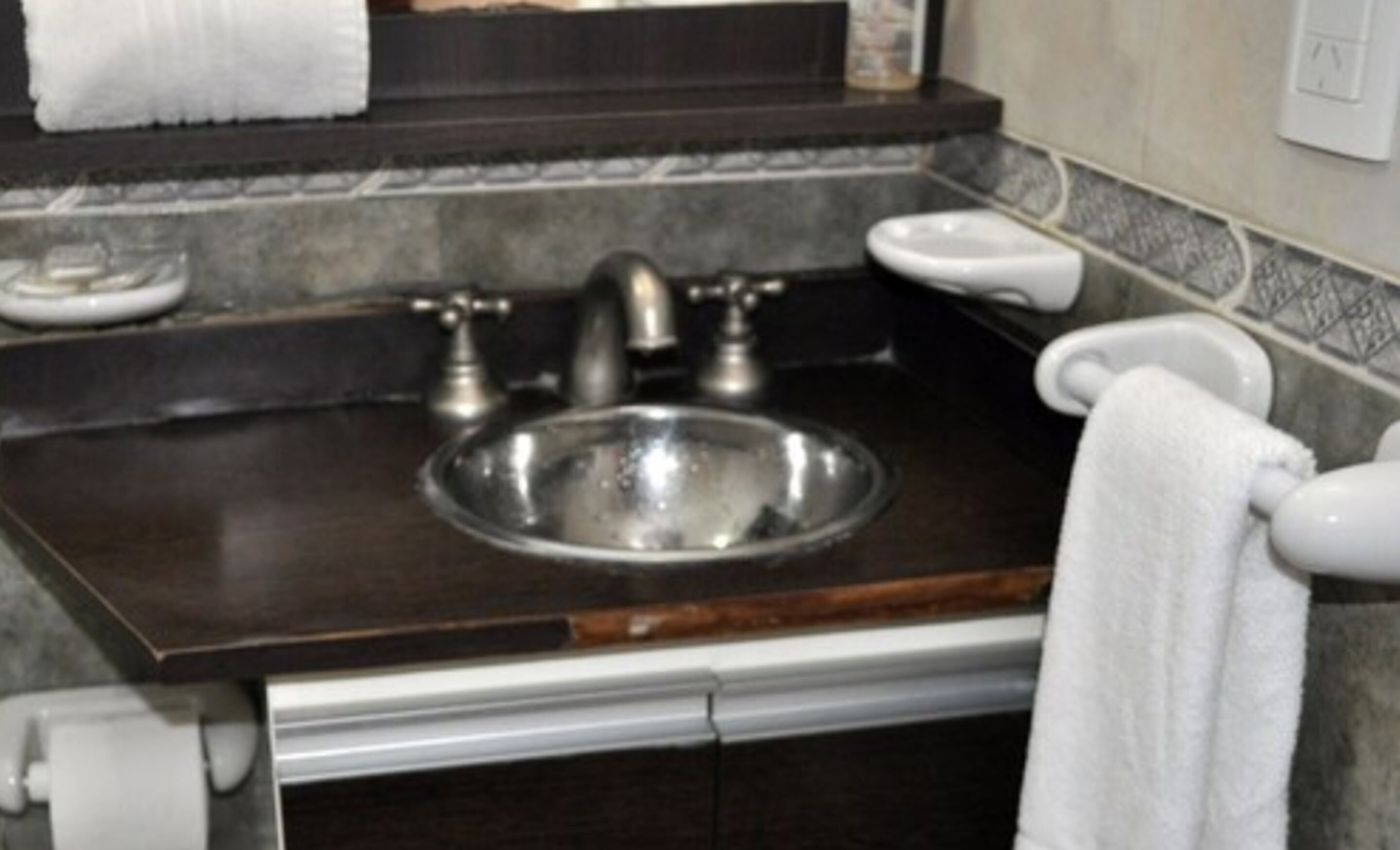 Une salle de bain avec un plan de toilette foncé, un évier en acier inoxydable, un miroir et un porte-serviettes.