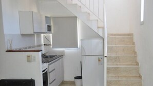 Cocina compacta con gabinetes blancos, un microondas y una estufa. Hay un cubo de basura y una escalera que sube a otro nivel.