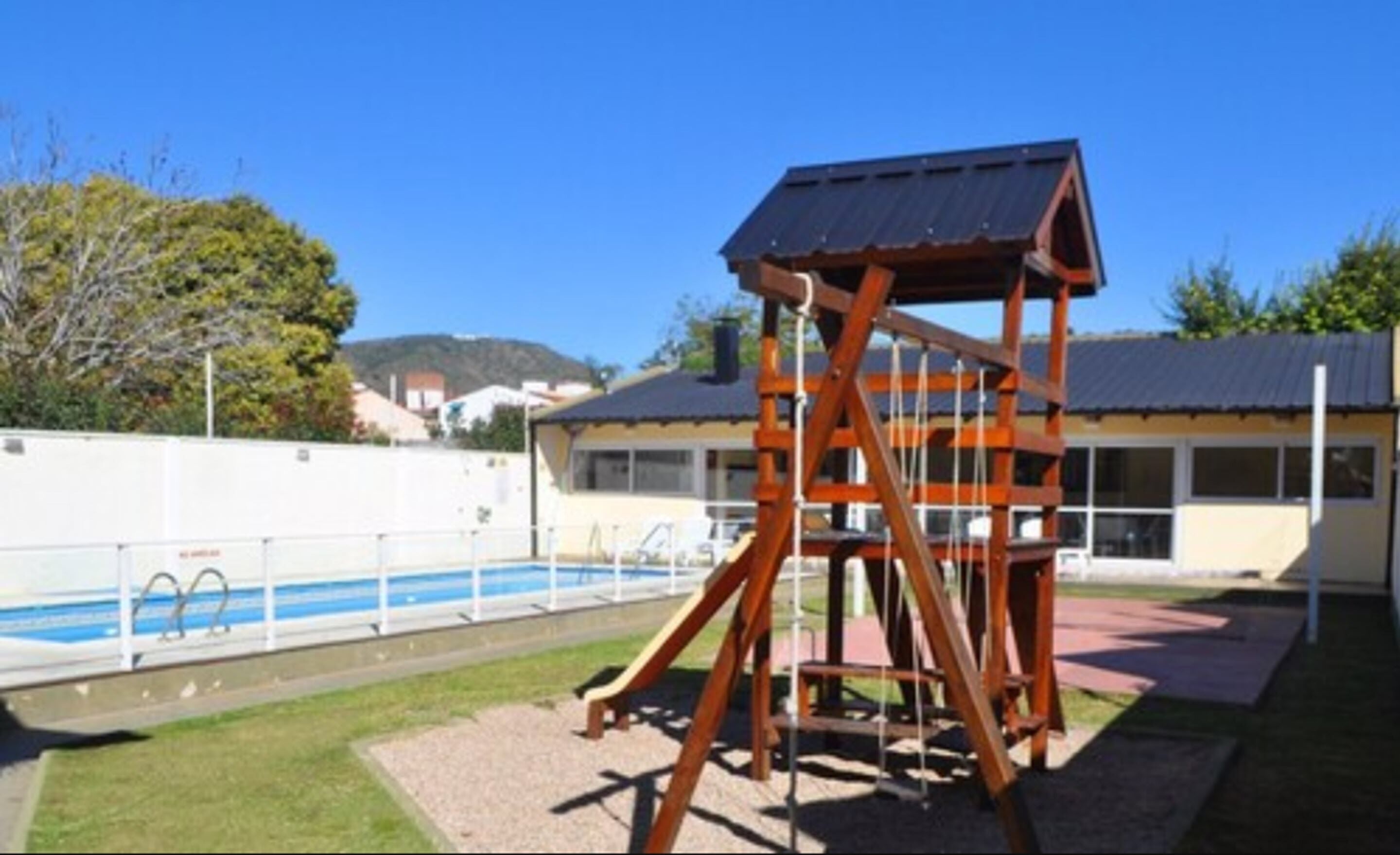 A playground with a swing set and a slide, a pool, and a building with a blue roof in the background.