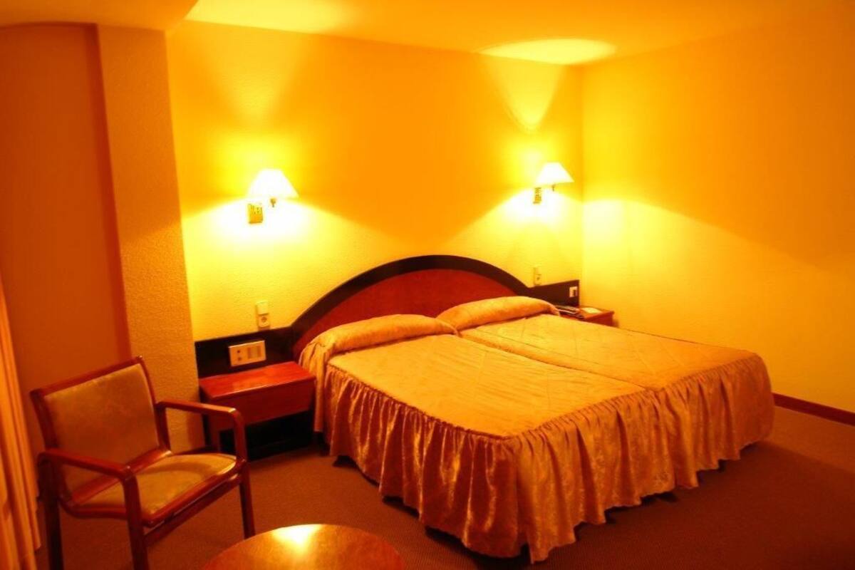 A hotel room with a double bed, two bedside lamps, a wooden chair, and a small table.