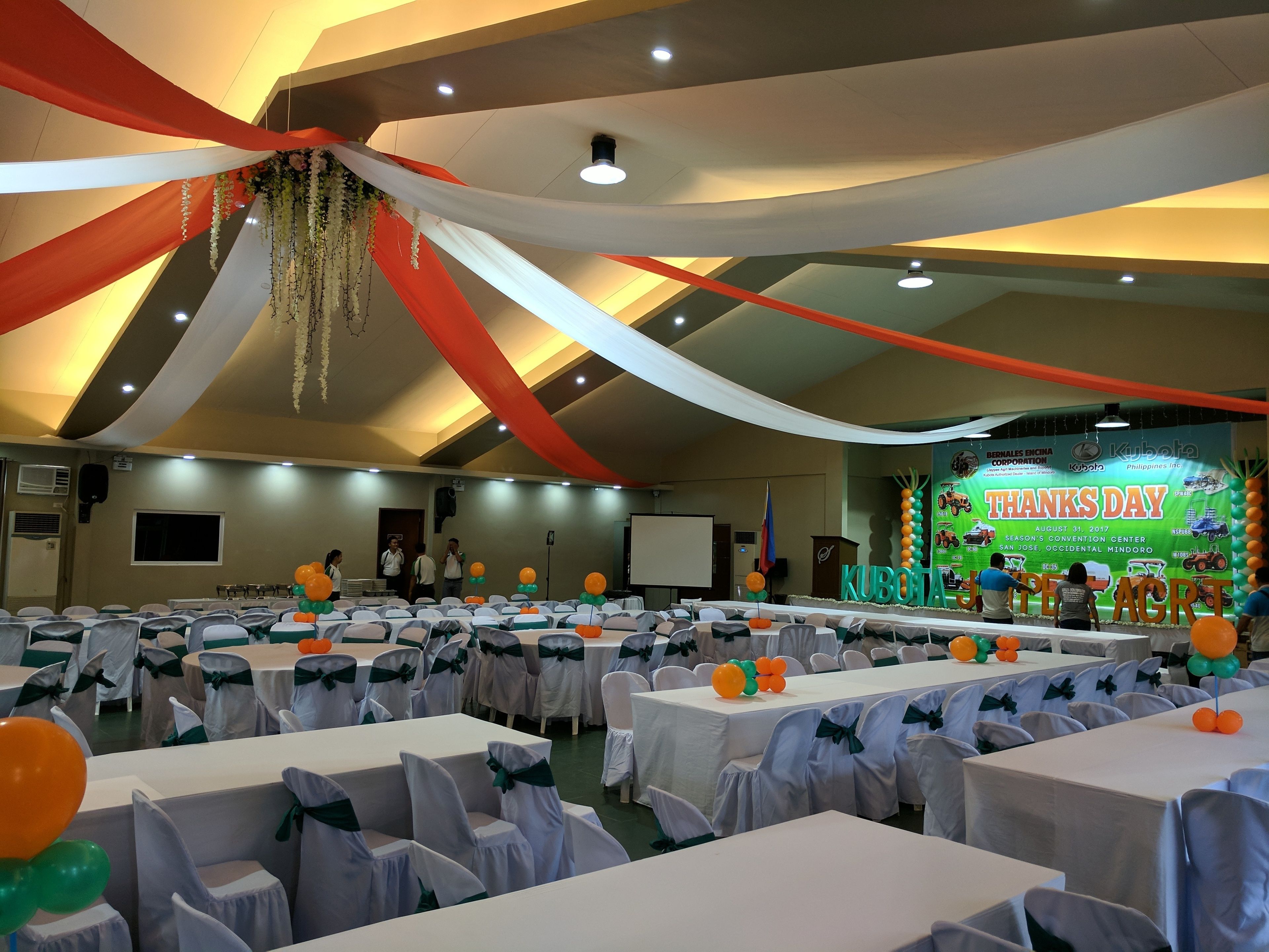 A banquet hall with tables covered in white tablecloths and chairs with green sashes, a stage with a 'THANKS DAY' backdrop, and a ceiling with draped fabric.