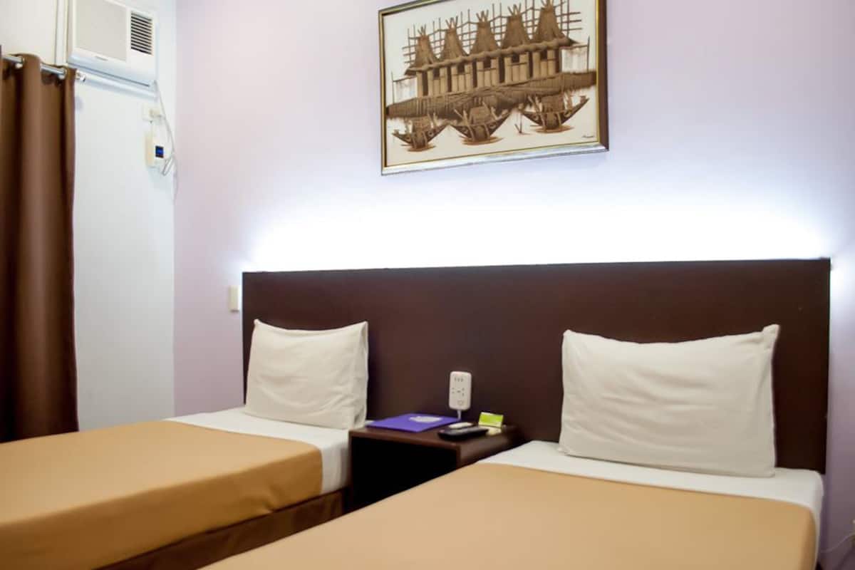 Ein Hotelzimmer mit zwei Einzelbetten, einem Nachttisch mit Lampe, einem gerahmten Bild an der Wand und einem Fenster mit Vorhängen.