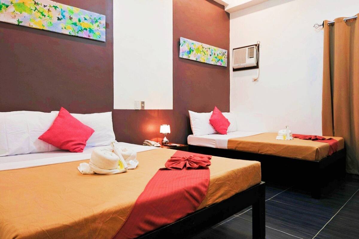 Ein Hotelzimmer mit zwei Betten, jedes mit einem roten Kissen und einer beige farbenen Tagesdecke. Ein kleiner Tisch mit einer Lampe und einer Wasserflasche steht daneben. Das Zimmer ist modern gestaltet, mit einer dunkelbraunen Akzentwand und einem farbenfrohen, abstrakten Gemälde über jedem Bett.