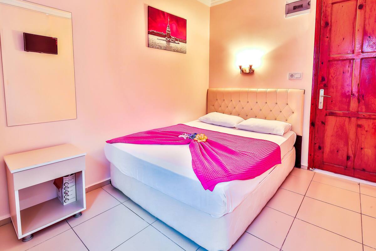 A neatly made bed with a pink blanket, a bedside table with a lamp, and a red door.