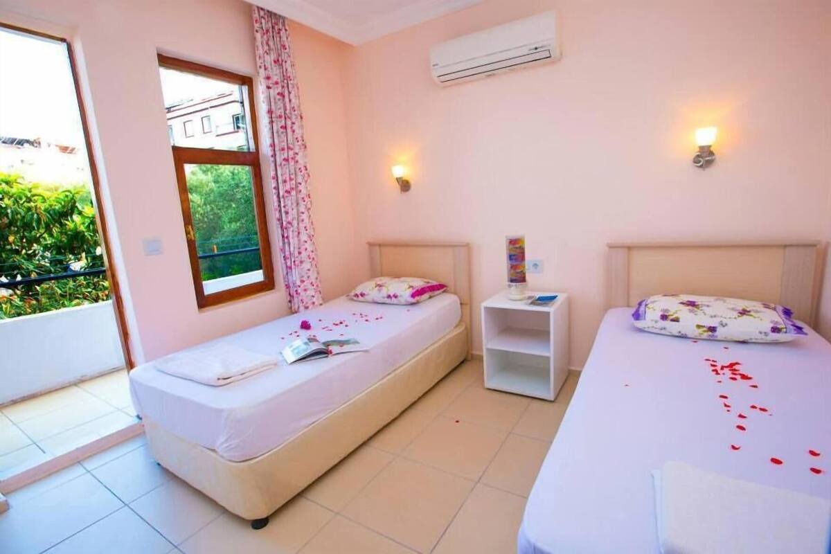 A bedroom with two beds, a window with curtains, an air conditioner, and a small bedside table.