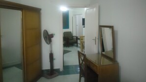 A hallway with a wooden door, a standing fan, a mirror, and a chair. - Jewel Mandara Apartments (Alexandria)