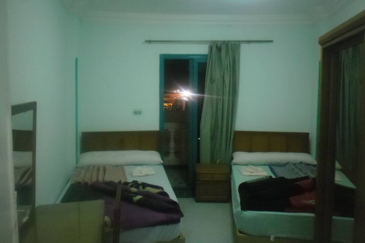 A small, dimly lit room with two single beds, a wooden nightstand, and a mirror on the left wall.