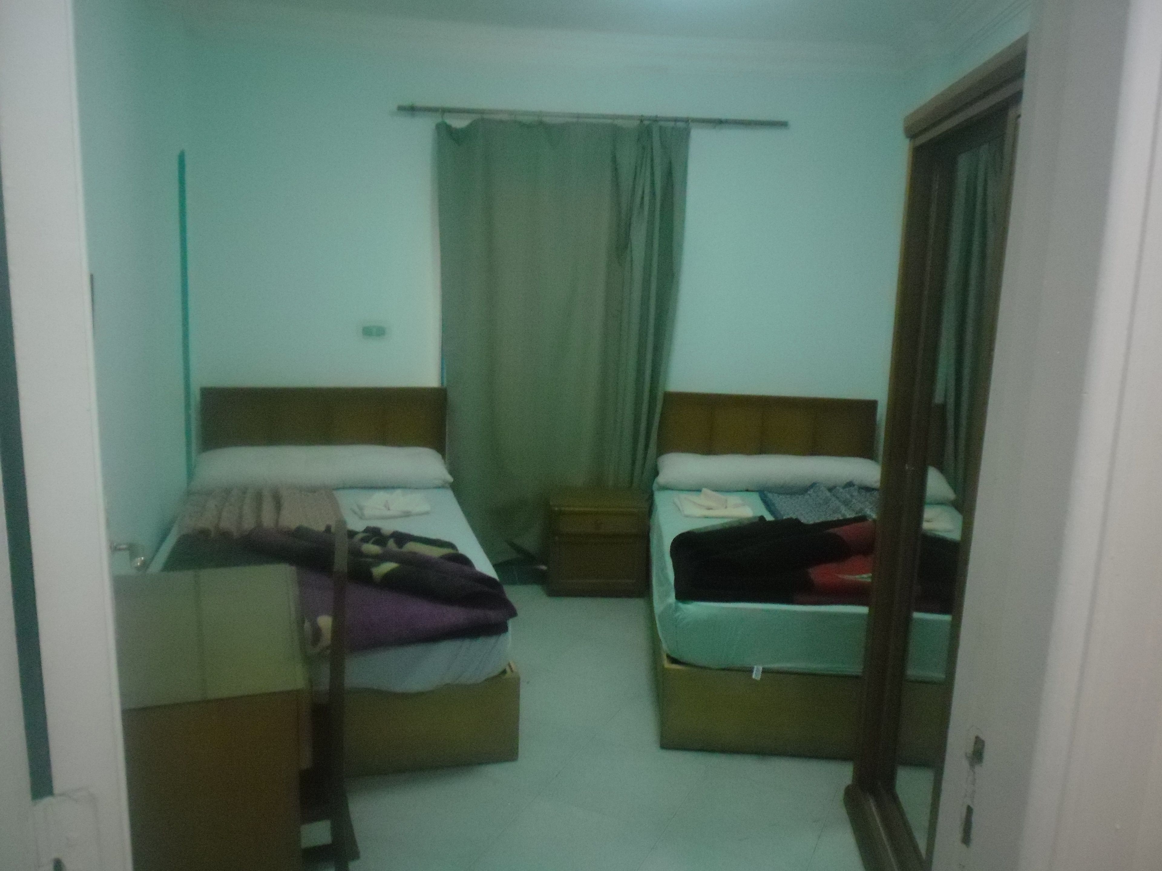A small, dimly lit room with two single beds, a wooden headboard, a small bedside table, and a curtain.