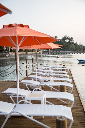 A wooden pier with white lounge chairs and orange umbrellas, overlooking a calm body of water with boats and a dock in the background. - Orcey Hotel (Datça)