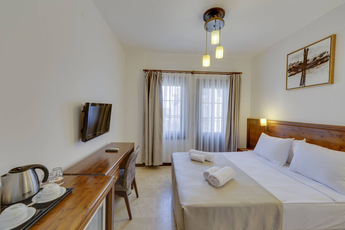 A hotel room with a bed, a desk with a kettle, a chair, a TV, and a painting on the wall.