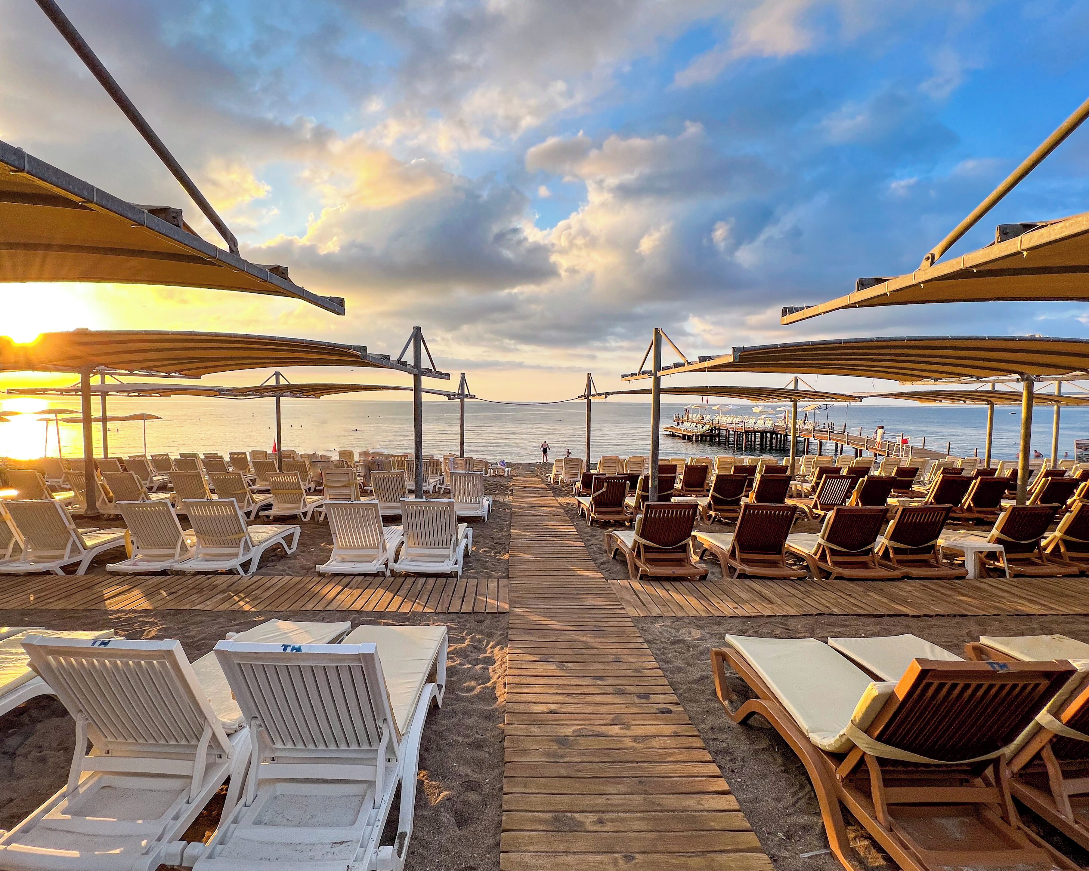 A beachside lounge area with rows of sun loungers under shaded canopies, a wooden walkway leading to the beach, and a pier in the background.