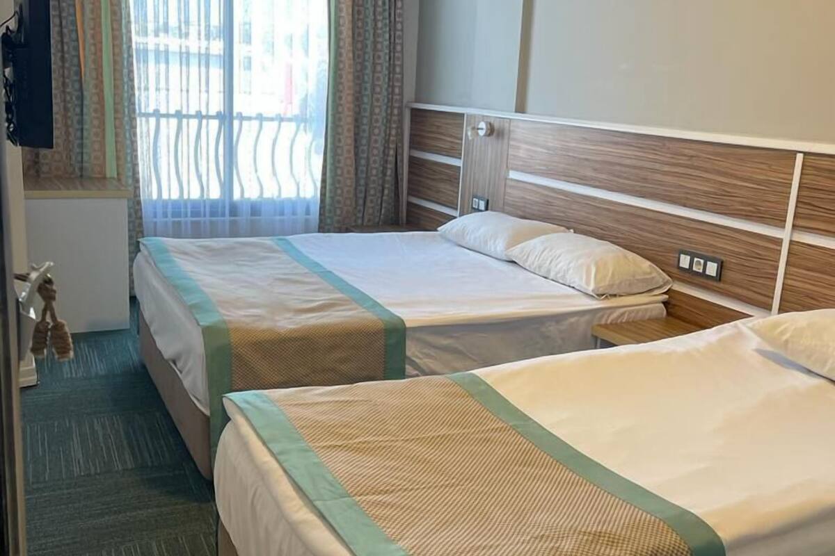 A hotel room with two beds, a window with curtains, an air conditioning unit, and a small table with a lamp.