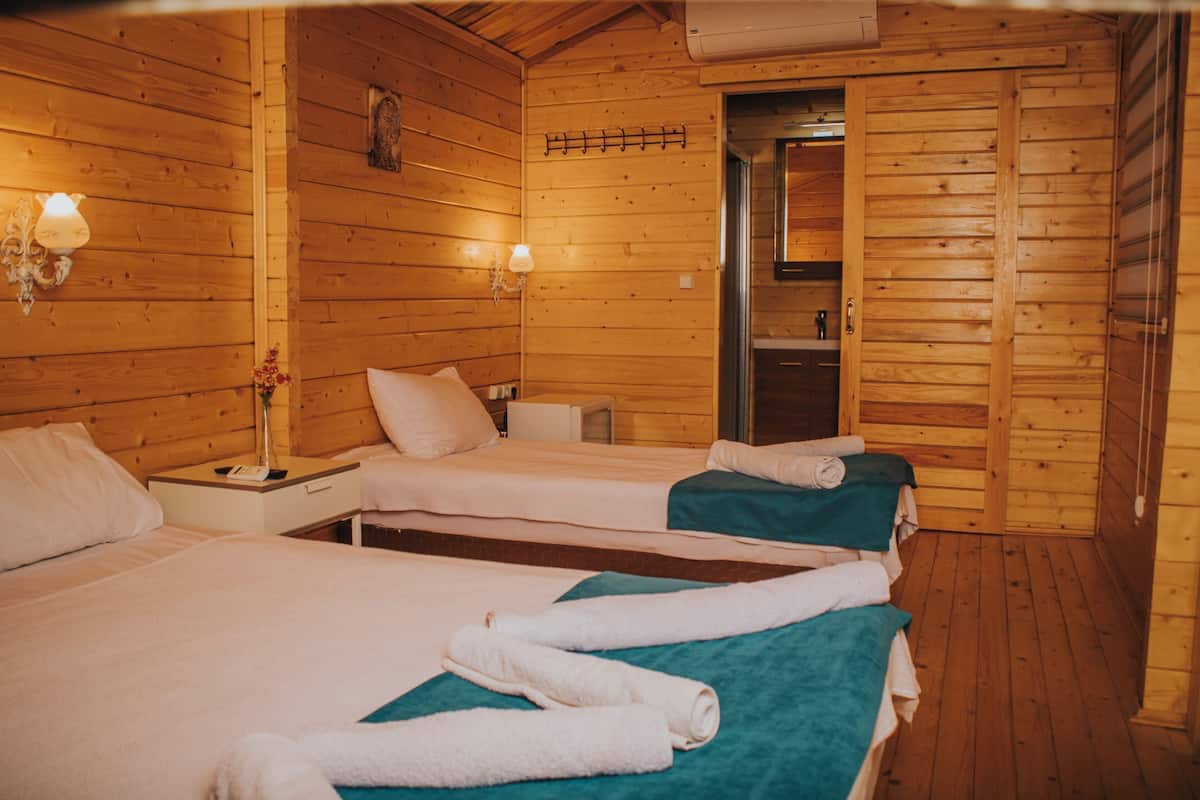 A wooden cabin-style room with two beds, a nightstand, a lamp, and a door leading to a bathroom.