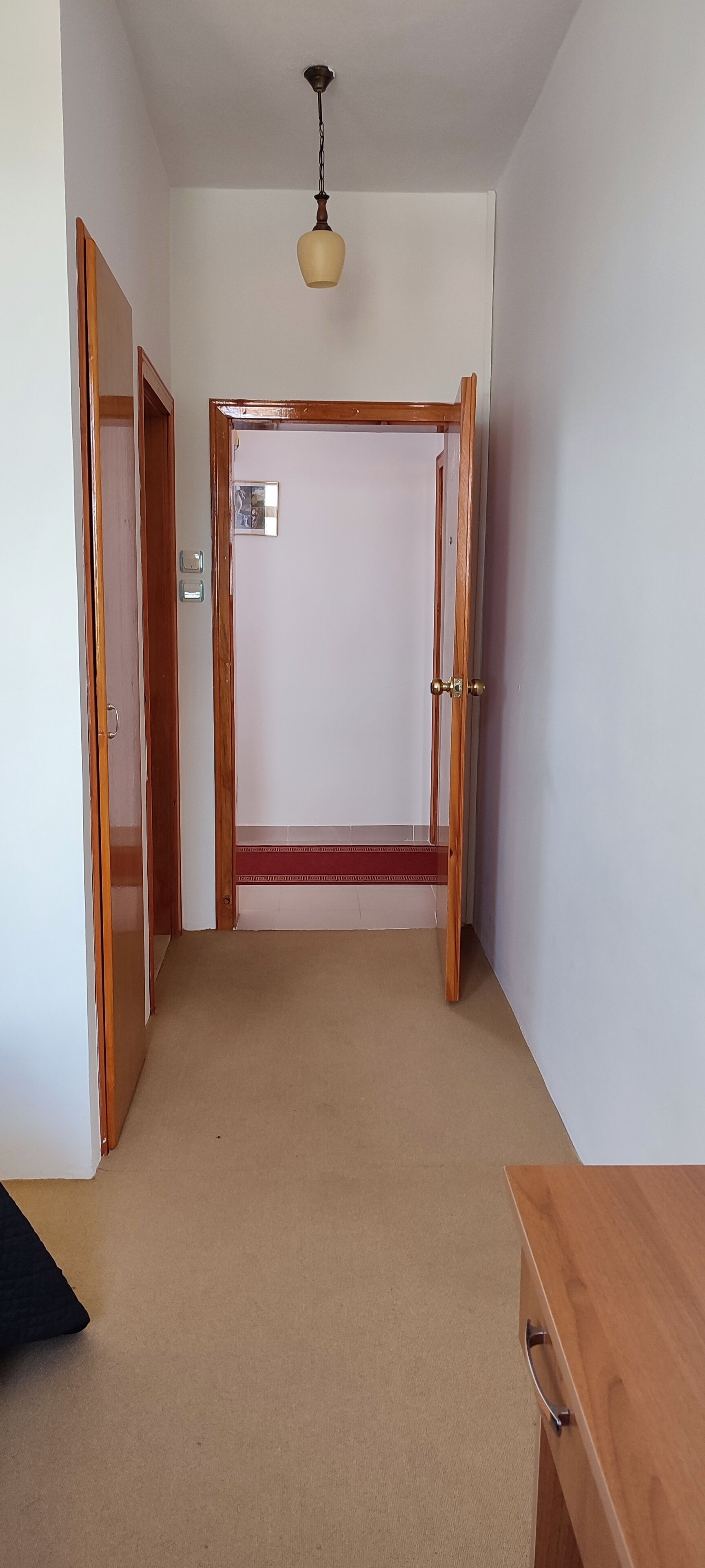 A narrow hallway with a wooden door at the end, a small light fixture above, and a wooden desk on the right.