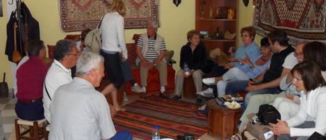 A group of people in a room with a large patterned rug, wooden furniture, and a shelf with decorative items.