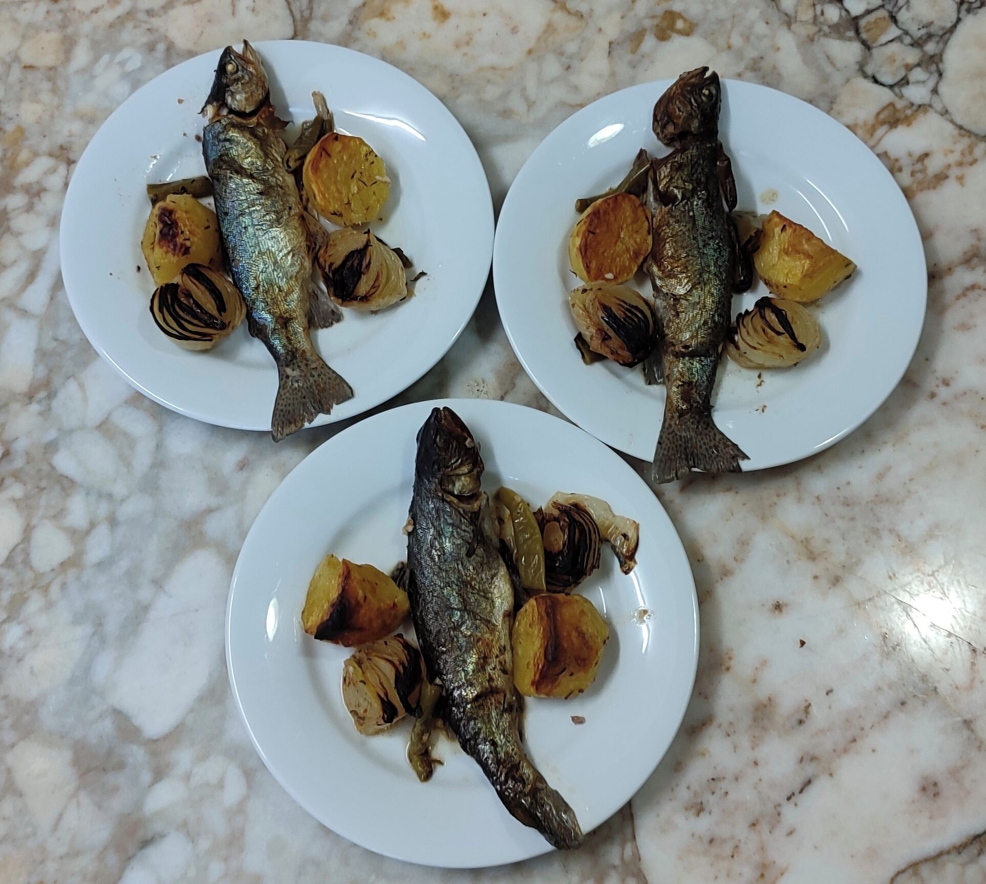 Tres platos de pescado a la parrilla y patatas asadas sobre una encimera de mármol.