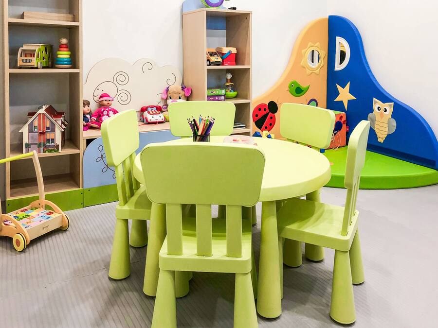 A children's play area with a round table and chairs, surrounded by shelves with toys and books.