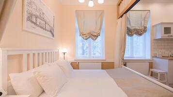A bedroom with a bed, a nightstand, a lamp, a window with curtains, a radiator, and a kitchen area with a microwave.