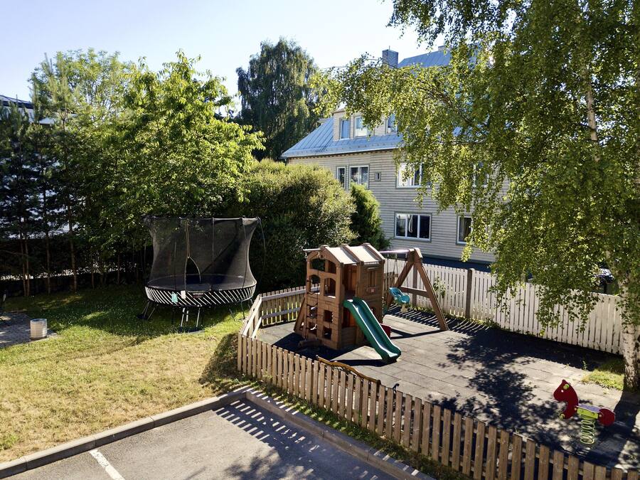 A playground with a trampoline, slide, and climbing structure, surrounded by a wooden fence and trees.