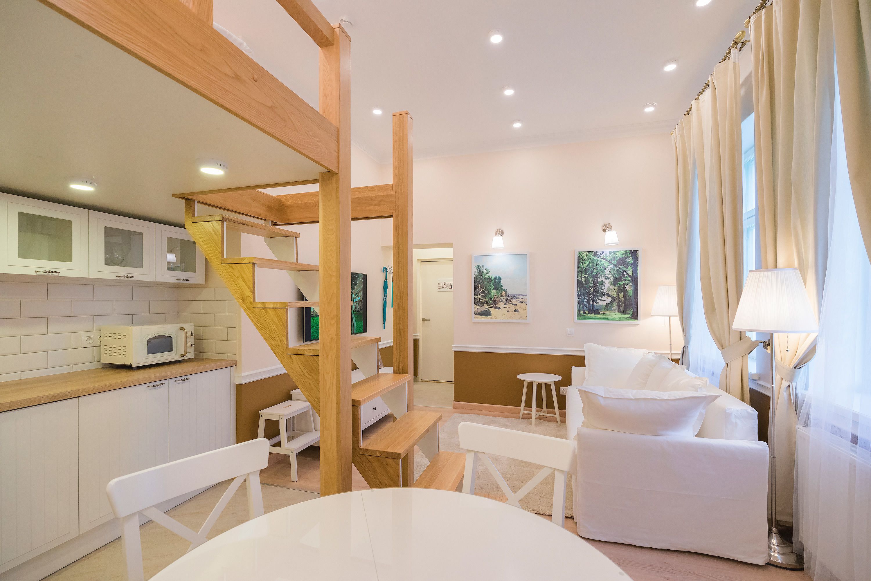 A modern kitchen with white cabinets, a microwave, and a wooden staircase. There is a dining area with a white table and chairs, and a white sofa.