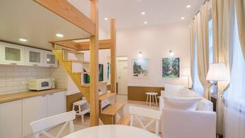 A modern kitchen with white cabinets, a microwave, and a wooden staircase. There is a dining area with a white table and chairs, and a white sofa.