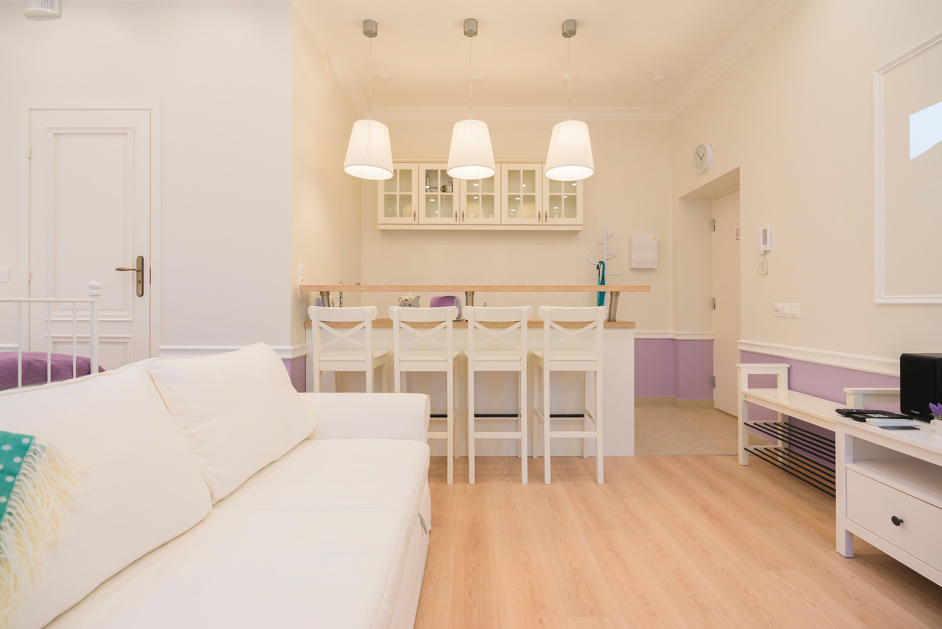A modern, well-lit room with a white sofa, a dining area with white chairs and a wooden table, and a small kitchen area with cabinets and a clock on the wall.