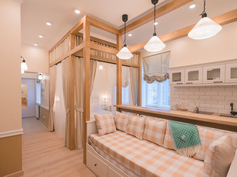A bedroom with a checkered bed, wooden ceiling beams, a window with curtains, a kitchen area with cabinets, and a hanging light fixture.