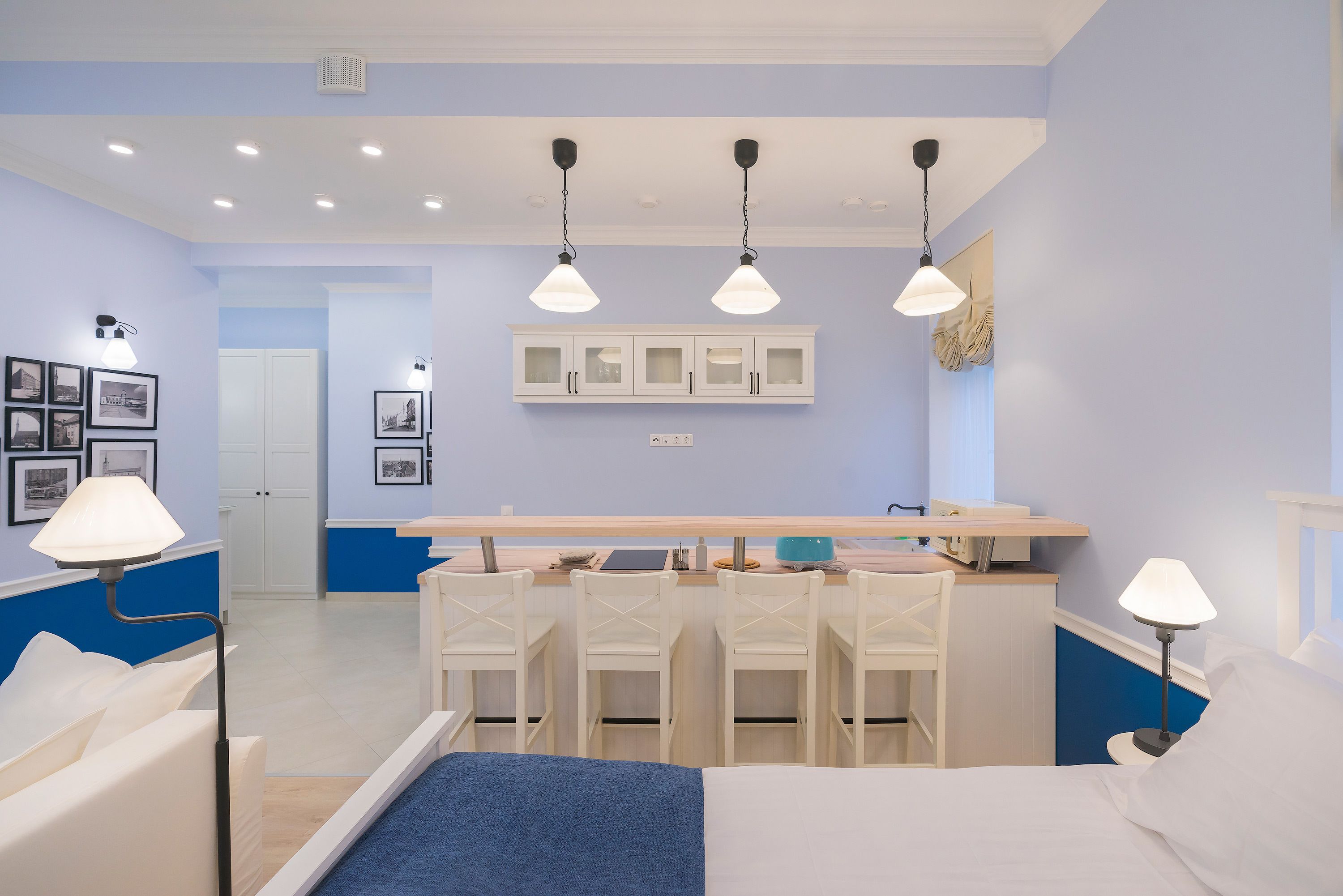 A bedroom with a bed, bedside tables, a desk, and chairs. There is a kitchenette with cabinets and a countertop.